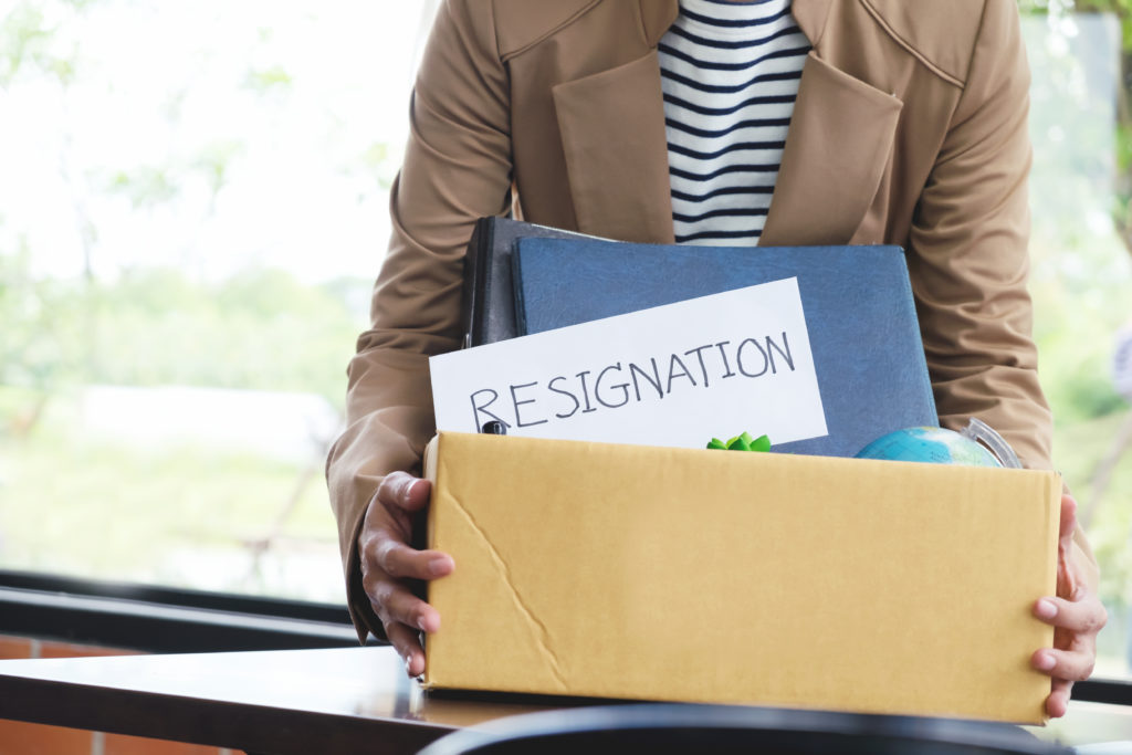 Employee exit survey help you understand why employees move on. Image shows a person holding a cardboard box full of belongings, with the word resignation written on it. 